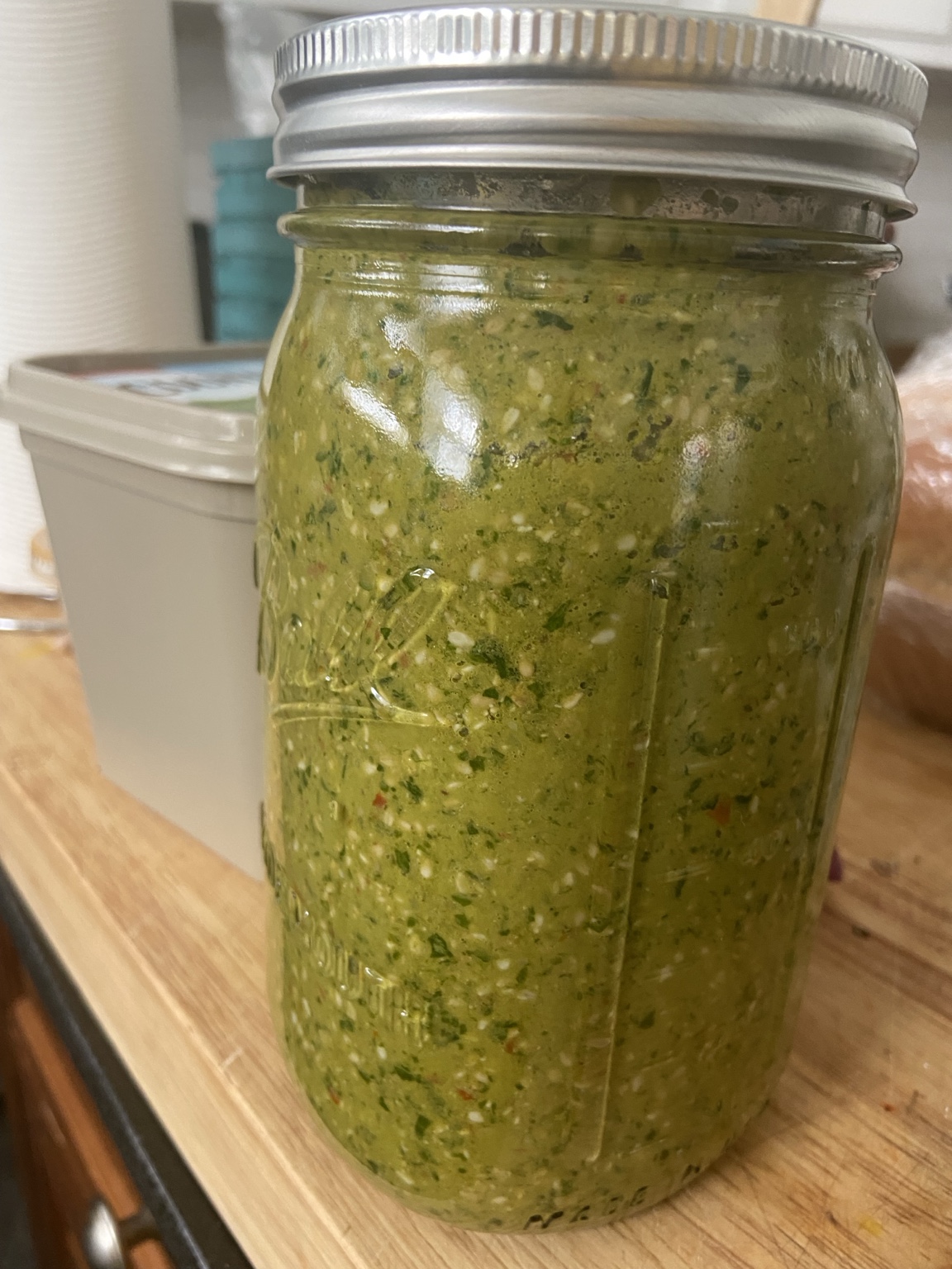 A tall mason jar filled with a homemade green herb blend flecked with red pepper, on a wooden counter
